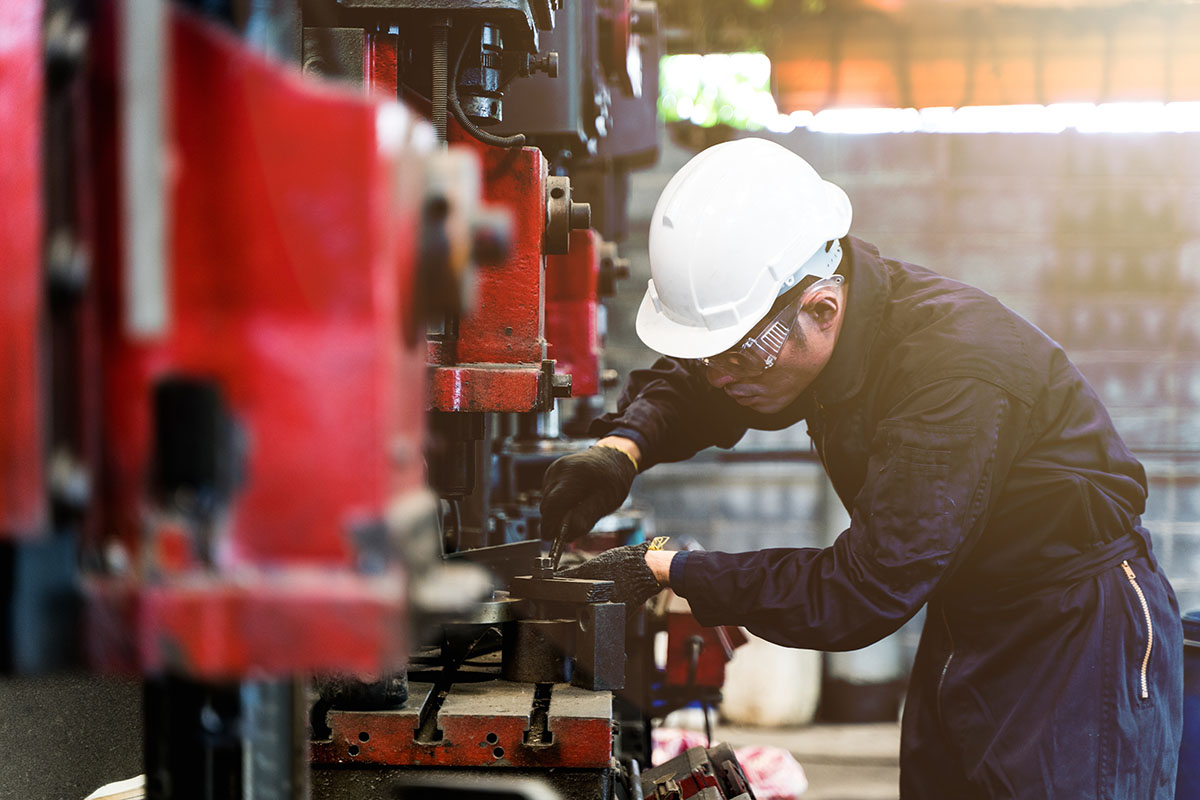 A maintenance technician in a white hard hat and safety glasses adjusting a large industrial press on a factory floor