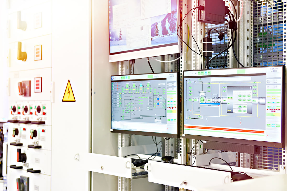 Two monitors showing process control diagrams mounted on an equipment rack beside an electrical switchgear panel